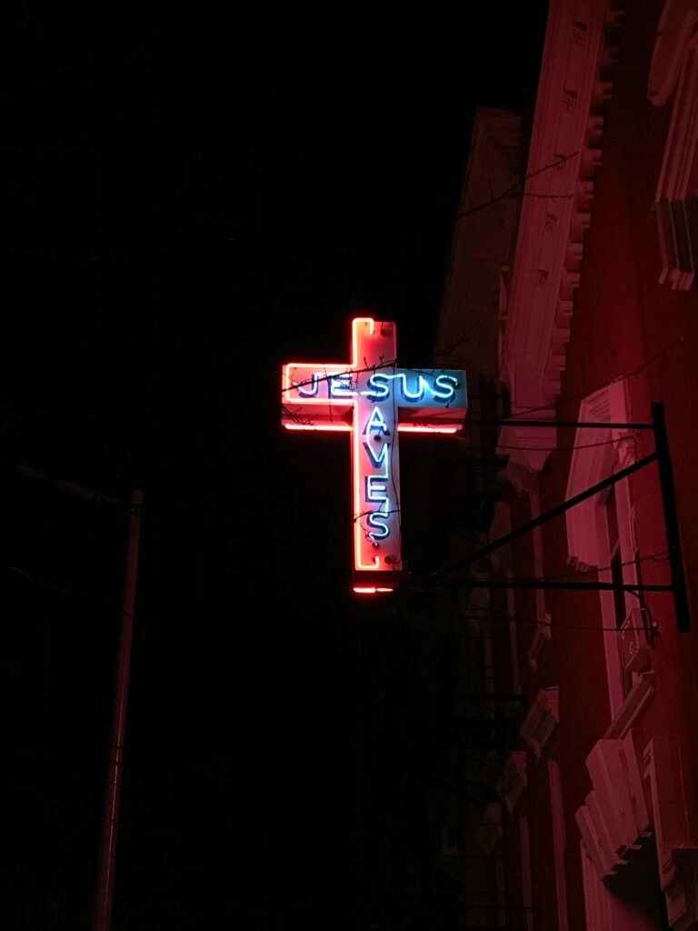 A glowing neon cross sign that reads 'Jesus Saves' against a dark night sky.